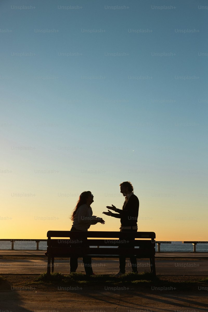 Silhouettes of two people talking on a bench at sunset ...